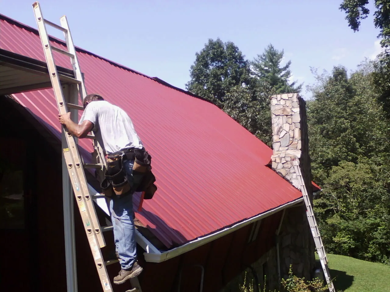 Roofer on a ladder beside a red metal roof and stone chimney — Soffit & Fascia Repair by Roofing Brothers in Midway